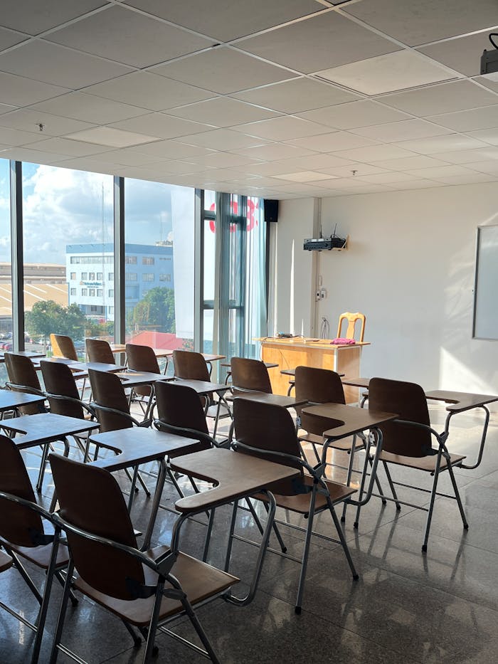 Bright and empty classroom with sunlight streaming through large windows, featuring wooden desks and chairs.